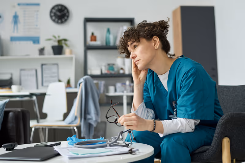 Female healthcare professional in teal scrubs sitting thoughtfully in a chair holding her glasses in a clinical office.