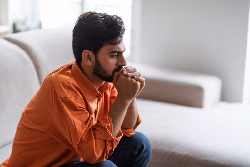 Thoughtful man in an orange shirt sitting on a couch with hands clasped near his face.