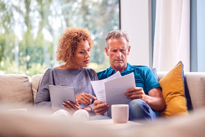 Middle-aged couple sitting on a sofa, reviewing documents together with a tablet and a coffee mug nearby.